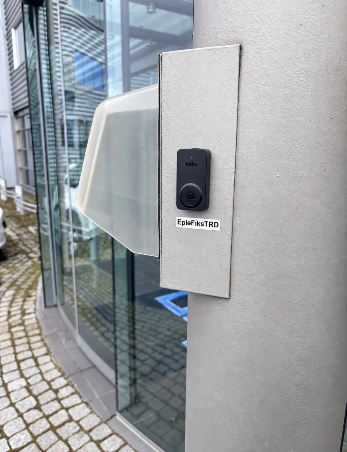 Svart dørklokke merket «EpleFiksTRD» på en grå søyle